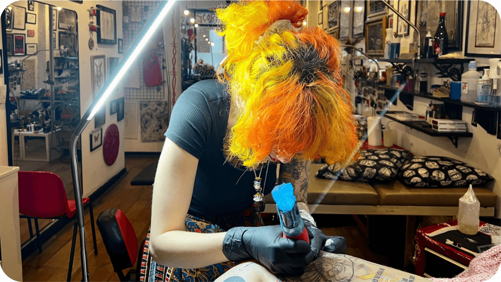  Vegan tattoo artist (Mogg) with brightly coloured hair leaning over and giving a tattoo to a client in a tattoo studio