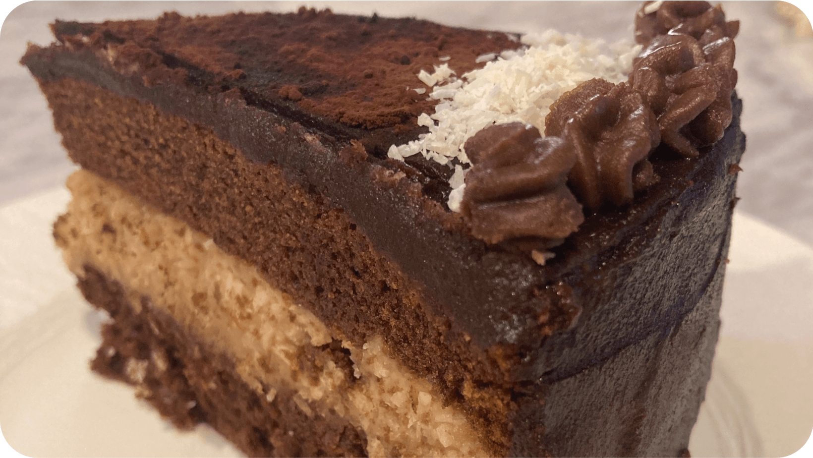 Big slice of layered vegan chocolate cake topped with coconut and chocolate cream on a white plate