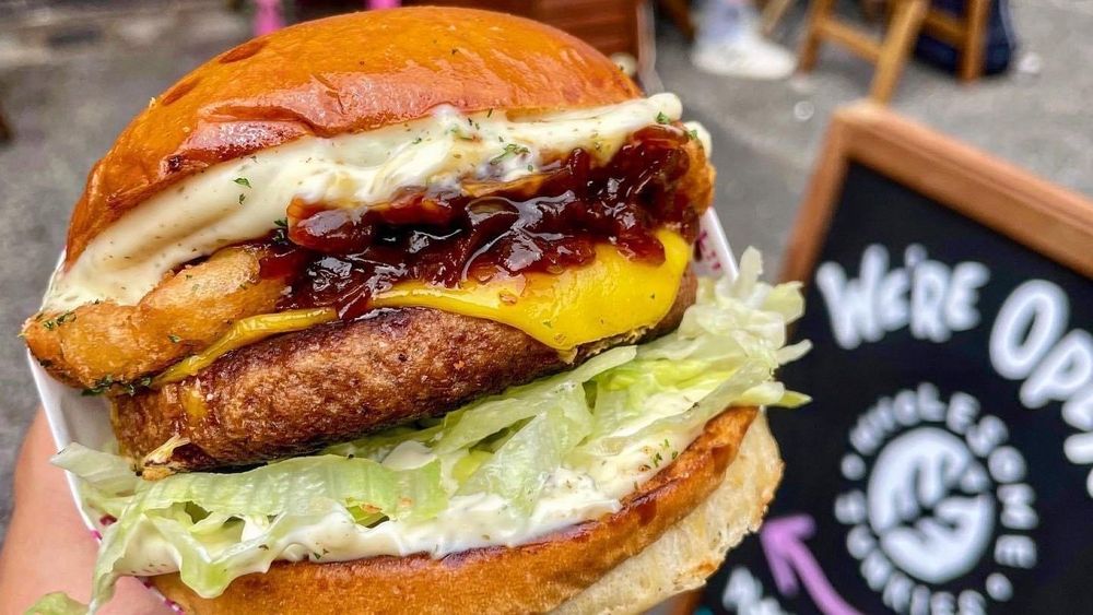 Vegan burger with vegan cheese, lettuce and onion rings in front of a Wholesome Junkies sign 