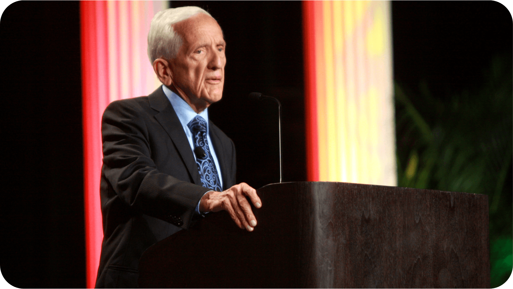 Image of elderly man with grey hair in a black suit (Dr T. Colin Campbell) giving a speech behind a lecturn