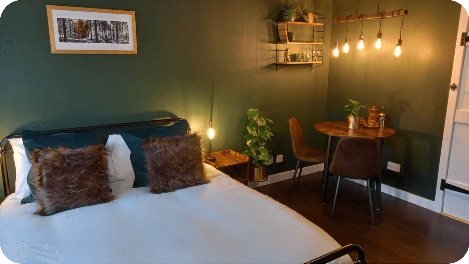 Interior of a cosy B&B room with a bed with pillows, wooden table and chairs, warm lighting and green walls