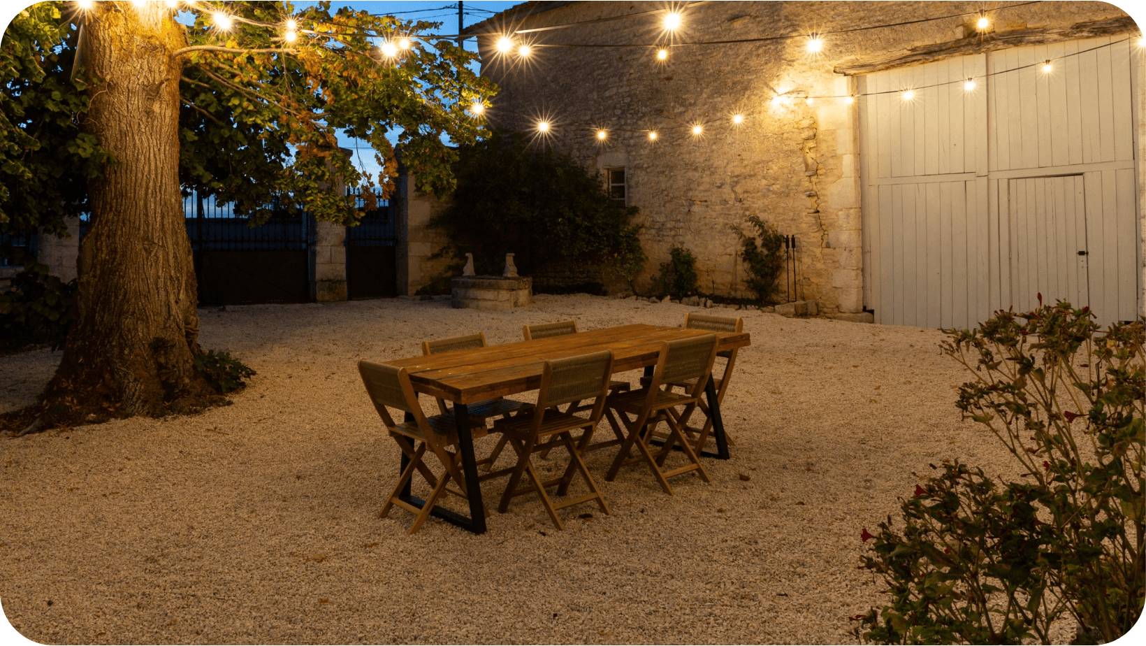 Outdoor wooden dining  table and chairs on gravel surrounded by lights and trees