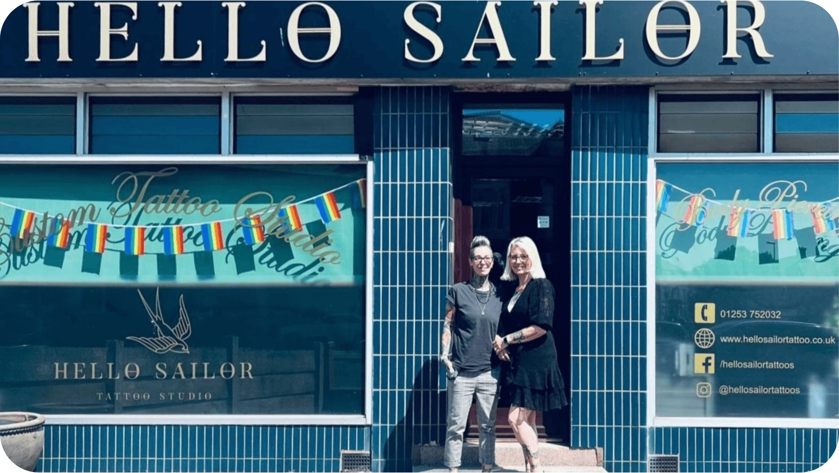 Two smiling women in front of their tattoo studio (Hello Sailor) which has Pride flags flying in the windows