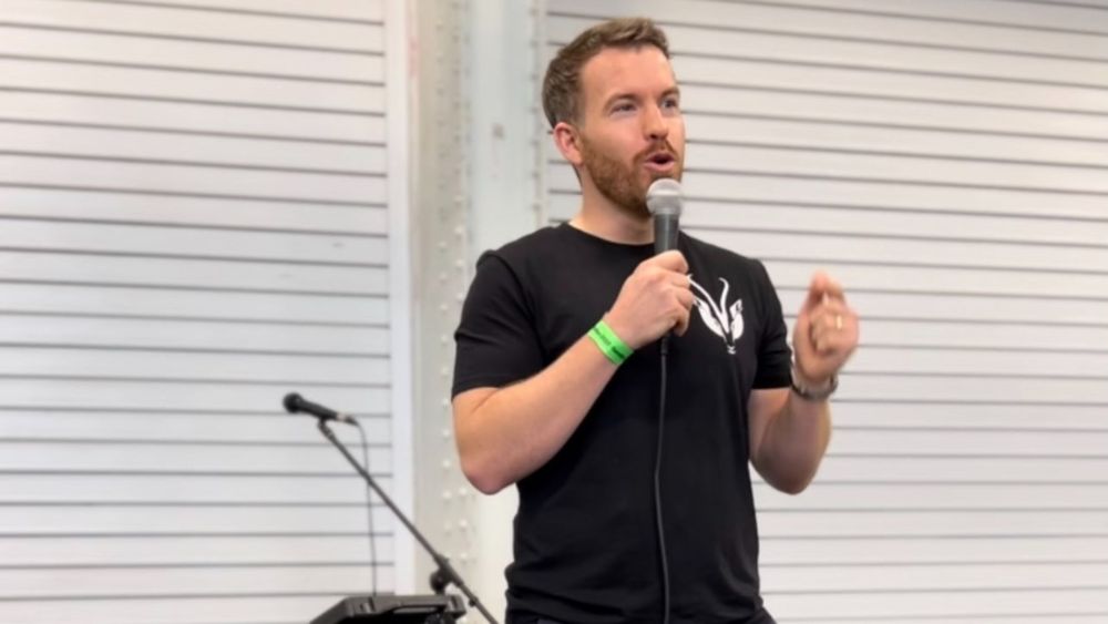 Man (Brendon Bale) in a black top talking into a microphone giving a speech in front of an audience