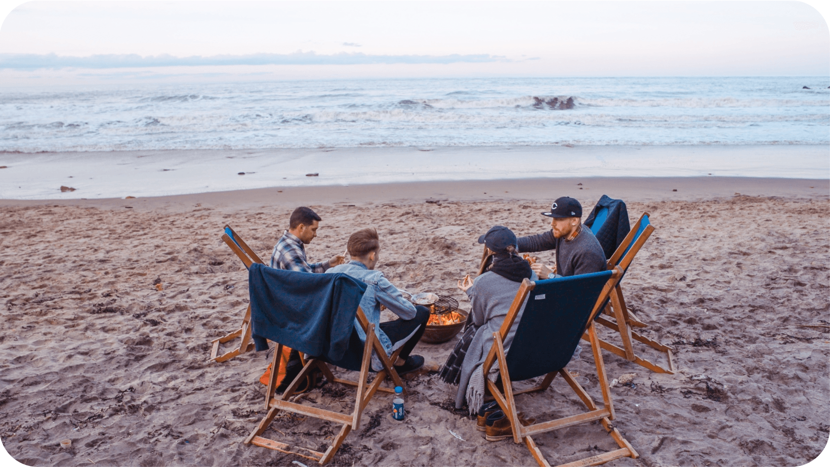  Group of friends sitting in deck chairs on the beach around a firepit