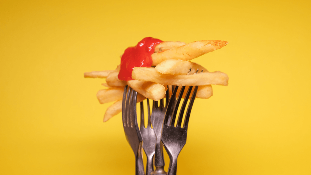 Chips covered with tomato ketchup being held up by forks on a yellow background.