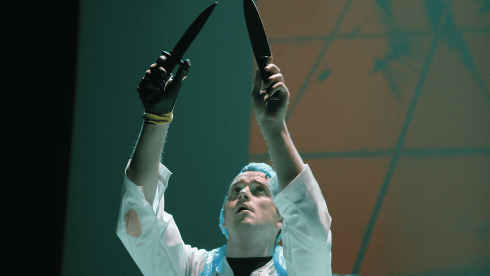 Slaughterhouse worker (from Blood On Your Hands play) holding two knives above his head and looking upwards