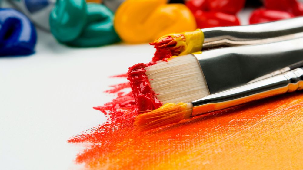 Paint brushes with paint on them on a partly painted canvas with multicoloured dollops of paints in the background