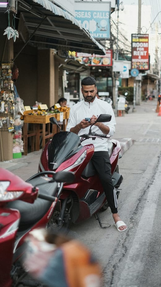 Man op een geparkeerde scooter kijkt op zijn telefoon, met een bord voor internationaal bellen erachter