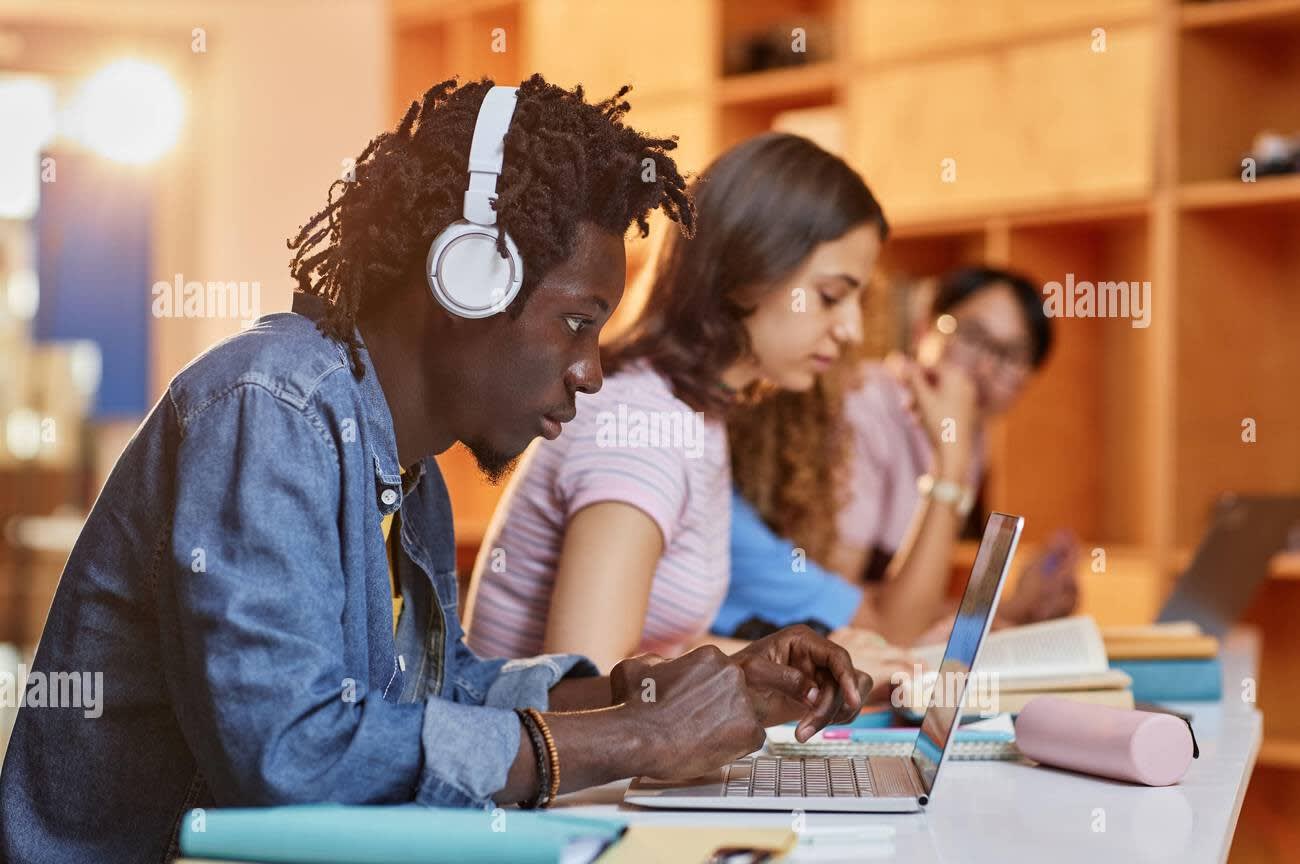 diverse group of students in row using laptops and studying in college library focus on black young man wearing headphones in foreground 2JRC07B