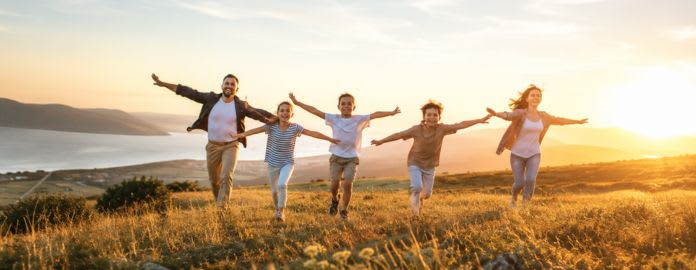 Famille avec trois enfants courant les bras ouverts dans une prairie au coucher du soleil, avec vue sur la mer et les collines - Vacances Bleues