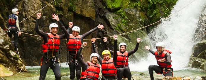 Groupe de personnes pratiquant le canyoning posant pour la photo les bras en l'air devant une cascade les pieds dans la rivière