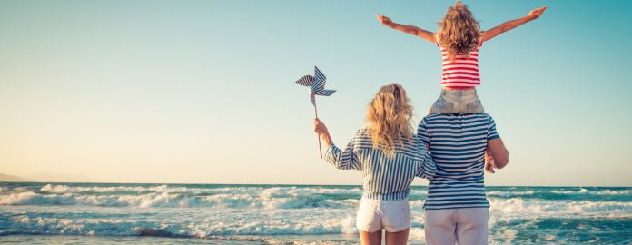Une famille de dos face à la mer agitée sur une plage ensoleillée. La mère tient un moulin à vent en papier et le père porte sa fille sur ses épaules, vêtus de vêtements à rayures de style marinière.