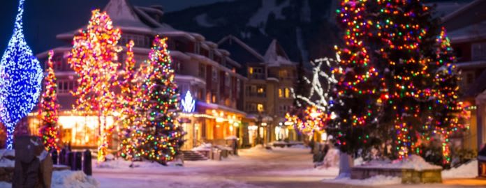 Photographie de nuit d'un village de ski alpin sous la neige, avec de nombreux sapins et chalets décorés de lumières de Noël multicolores et scintillantes