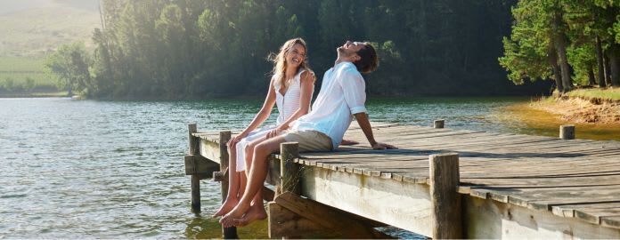 Couple souriant assis sur un ponton en bois sur un lac en campagne - Vacances Bleues