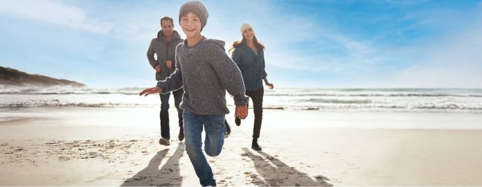 Enfant souriant courant en premier sur la plage avec ses parents derrière, sous un ciel bleu. Vacances Bleues