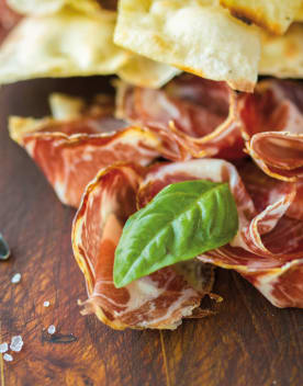 Tranches fines de charcuterie italienne avec du pain croquant, garnies d’une feuille de basilic frais, sur une planche en bois.