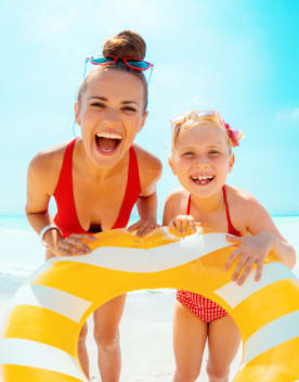 Mère & fille sur la plage en maillot de bain rouge tenant une bouée rayée jaune et blanche sous un ciel bleu, la mer en arrière plan - Vacances Bleues