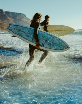 Deux surfeurs courant dans les vagues avec leur planche sous le bras - Vacances Bleues
