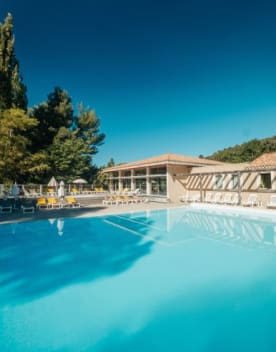 Grande piscine extérieure ensoleillée avec transats et parasols, adossée à un bâtiment à toit de tuiles et une forêt de pins sous un ciel bleu clair. Domaine de Château Laval - Gréoux les Bains
