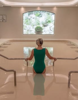 Femme de dos portant un maillot de bain une pièce vert canard dans un bassin de spa élégant avec une vue sur le jardin - Château Laval / Vacances Bleues