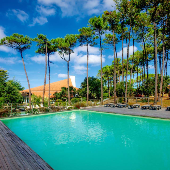 Piscine extérieure turquoise avec terrasse en bois, chaises longues, entourée d'une pinède sous un ciel bleu avec nuages - Domaine de l'Agréou Seignosse