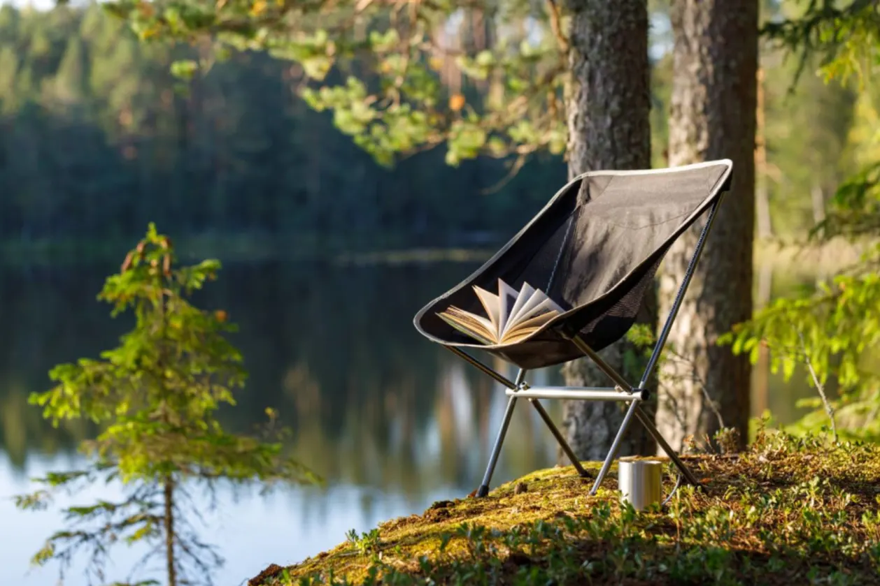 camping folding chair with book and mug with forest lake background