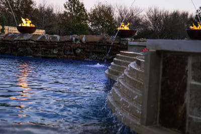 Wide view of a resort-style pool in Mansfield, TX featuring a three-structure grotto with cascading waterfalls and six copper fire bowls reflecting across the water surface.