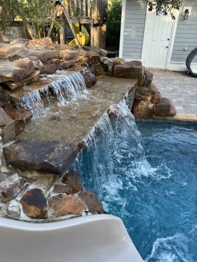 Waterfall cascading from a raised deck into a backyard pool with blue water at the Colon Residence in Santa Rosa Beach, FL.