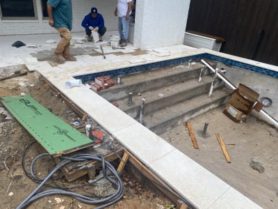 Geometric pool under construction in Dallas, TX, revealing gunite shell, multi-depth entry steps, travertine coping, and ledgestone-veneered retaining walls with integrated staircase.