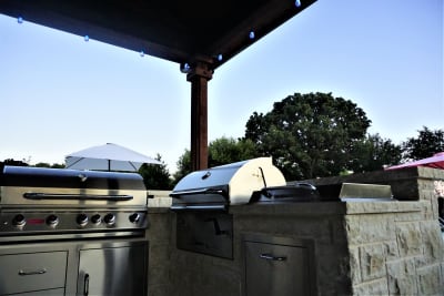 Establishing view of an outdoor kitchen with Bull refrigerator, natural gas grill, and raised beam structure on Leudar Buff coping at a Tarrant County estate in Arlington, TX.