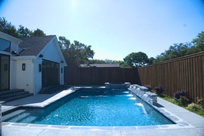The raised spa's Burlington quartzite classico coping creates clean lines while multiple sheer descents cascade into the pool below, photographed in natural daylight.