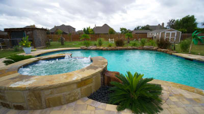 Establishing overview of a family resort pool with stone coping, tiered spillover, and weeping wall water feature at a Haslet, Tarrant County, TX residence.