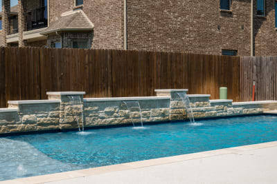 Material-detail view of Arctic White pebble plaster pool surface alongside Buff Lueders stone veneer and March Wind spray deck finish in North Richland Hills, Tarrant County, TX.