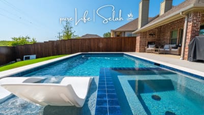 Establishing overhead view of a geometric pool under construction in Roanoke, Denton County, TX, revealing the gunite shell, tanning ledge, spa bench, spa step, bubblers, and spray deck.