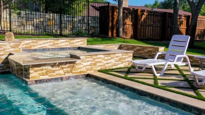 A family resort pool in Irving, Dallas County, TX featuring a waterfall, pool slide, love seats, travertine and granite coping, and a tile-veneered custom spa on a stone and paver deck.