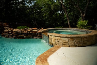 Establishing view of a naturalistic freeform spa with Noble RF195 Zaffre tile, Oklahoma stone fireplace with wood storage, and weeping wall spray deck at a private residence in Arlington, TX.