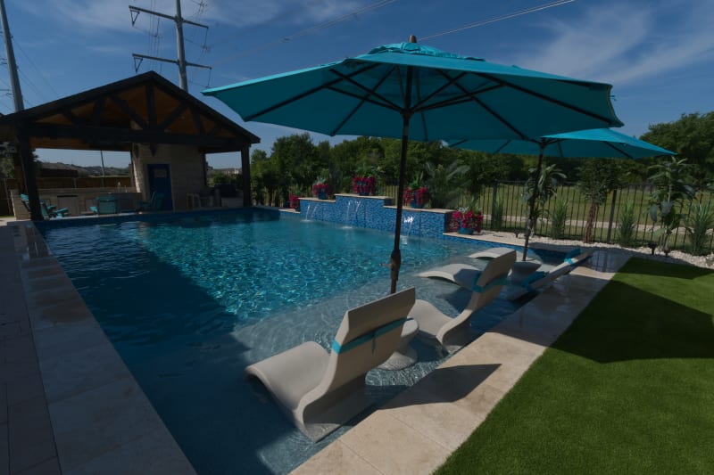 Architectural pool entry featuring tanning ledge at 18-inch depth with integrated umbrella sleeve for shade. Regatta blue pebble finish complements peacock tile raised beam walls and French pattern stone pavers.