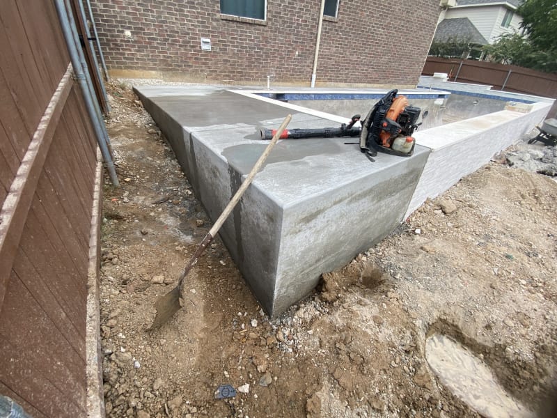 Mid-construction view of a geometric pool taking shape in Hurst, Tarrant County, TX, revealing the gunite shell, tanning ledge formation, and retaining wall structure before Caribbean Blue Peerless Pebble finish is applied.