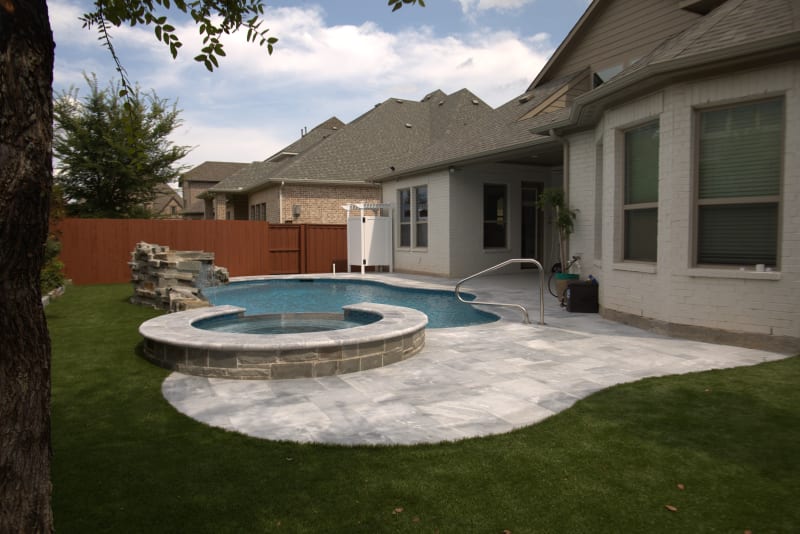 Establishing overview of a naturalistic freeform pool with Silver Mist grotto, Burlington Armani Grey coping, and Peerless Pebble Blue Ice Harbor finish in Frisco, Denton County, TX.