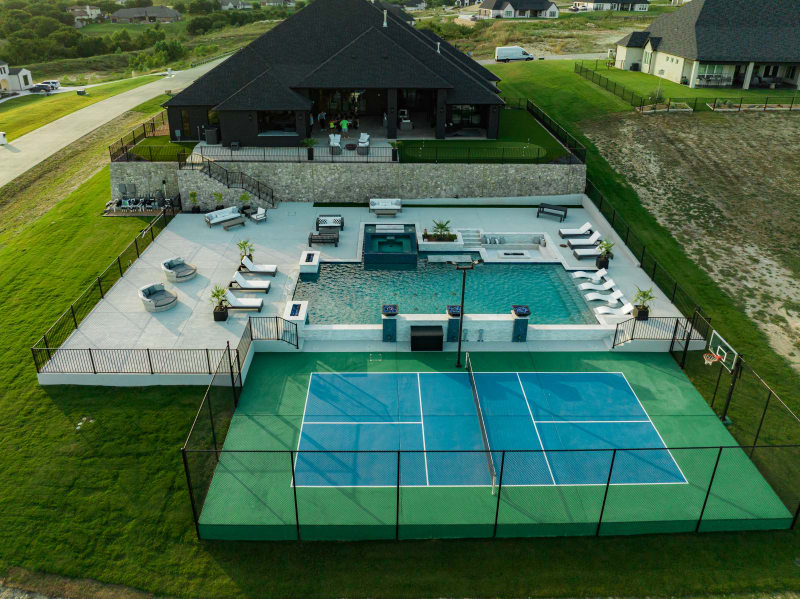 Drone aerial of the complete Rodgers property in Fort Worth showing the geometric pool, raised spa, sunken fire pit, copper fire bowls, sport court, and landscaping