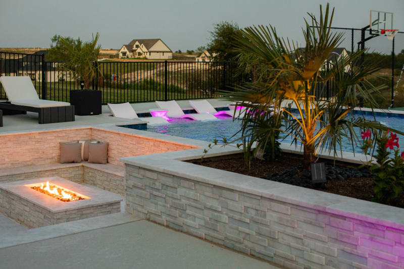 Sunken fire pit with active flame at dusk, tanning ledge with purple LED-lit in-pool loungers in the background