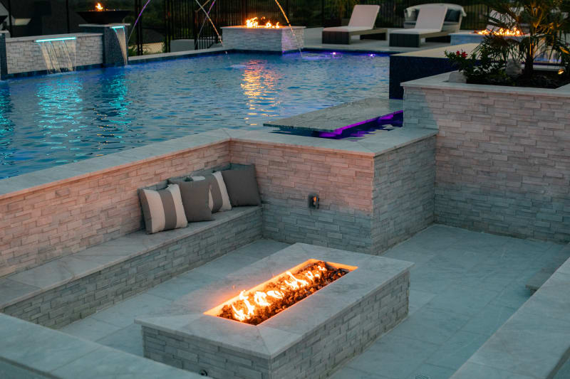 Twilight view from the sunken fire pit across the pool — fire pit glowing, copper fire bowls lit, LED bubblers and sheer descents active, laminar jets arcing over the water