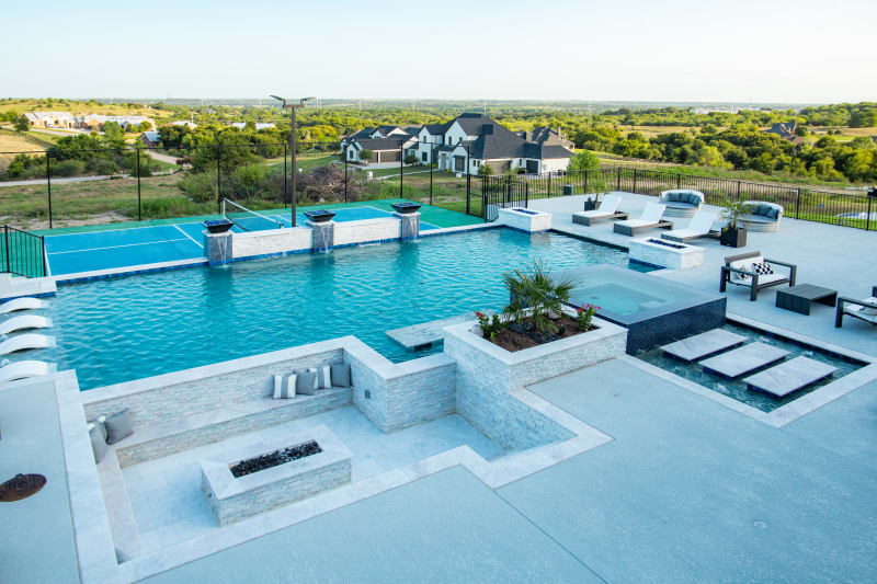 Wide view of the Rodgers outdoor environment from the covered patio — geometric pool with sheer descents cascading along the raised beam wall, copper fire bowls, raised spa, sunken fire pit area, and sport court beyond, framed by the Fort Worth landscape
