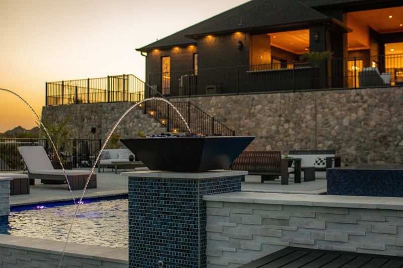 Copper fire bowl with laminar water jets arcing into the pool at sunset, with the house glowing warm in the background — Rodgers outdoor environment in Fort Worth by Selah Pools
