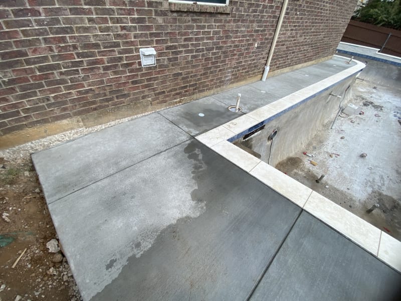 Mid-construction view of a geometric pool shell in Hurst, TX, revealing the structural foundation for three 18-inch sheer descents and a negative-edge roll bond beam against a brick retaining wall.