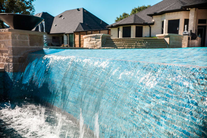 Close-up of the negative edge with water flowing over Aquabella Bora Bora 647 glass tile in 1x2 format, Buff Lueders stone columns visible behind, at the Sharp residence in Granbury, TX
