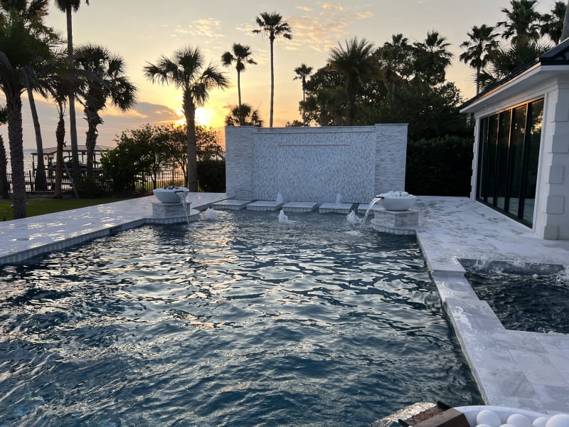 Four fire and water bowls ignite above a Blue Surf plaster pool with French pattern marble pavers and travertine ledgestone walls at a Miramar Beach, FL coastal retreat.