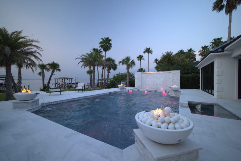 Hero view of a custom resort-style pool in Miramar Beach, FL, featuring a raised fire feature, bubblers, 8-foot linear LED sheer descent, tanning ledge, and French pattern marble paver deck with travertine ledgestone veneer walls.