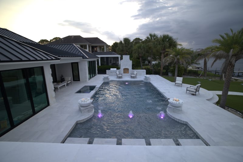 Establishing aerial overview of a custom resort-style pool with travertine ledgestone walls, French pattern marble pavers, and fire and water bowls at a Miramar Beach, FL coastal estate.